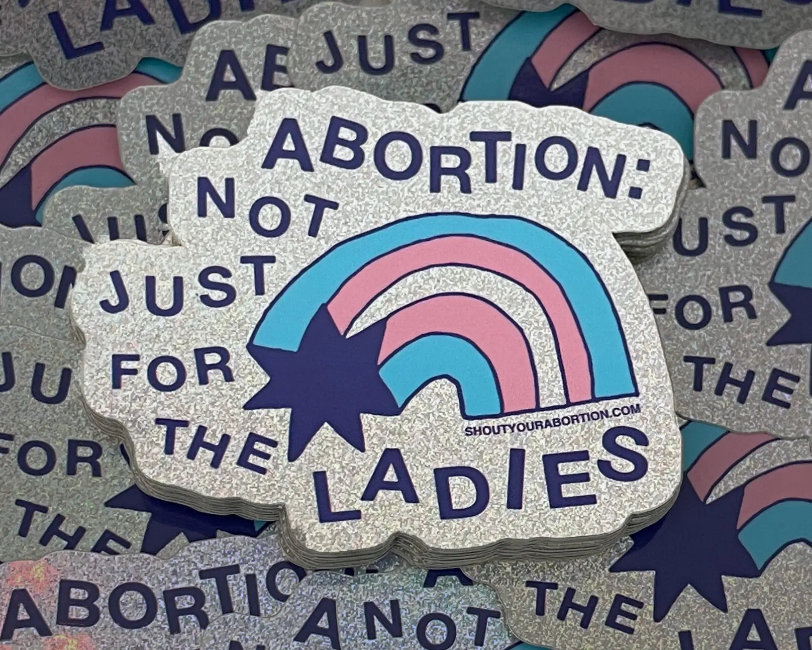 Holographic glitter sticker with a blue, pink and dark blue rainbow and the words ABORTION: NOT JUST FOR THE LADIES