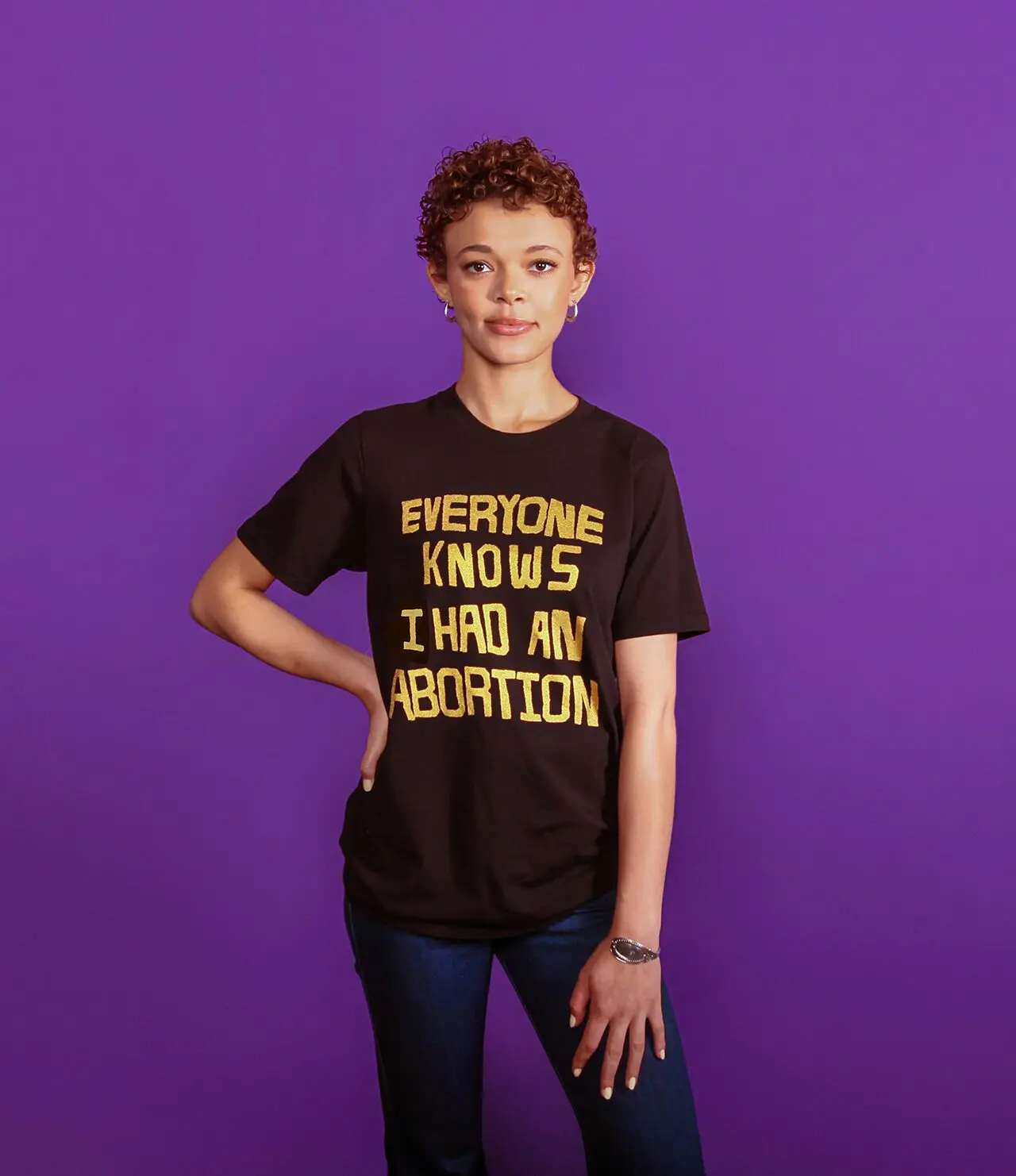 Model wearing a black tee that says EVERYONE KNOWS I HAD AN ABORTION in gold
