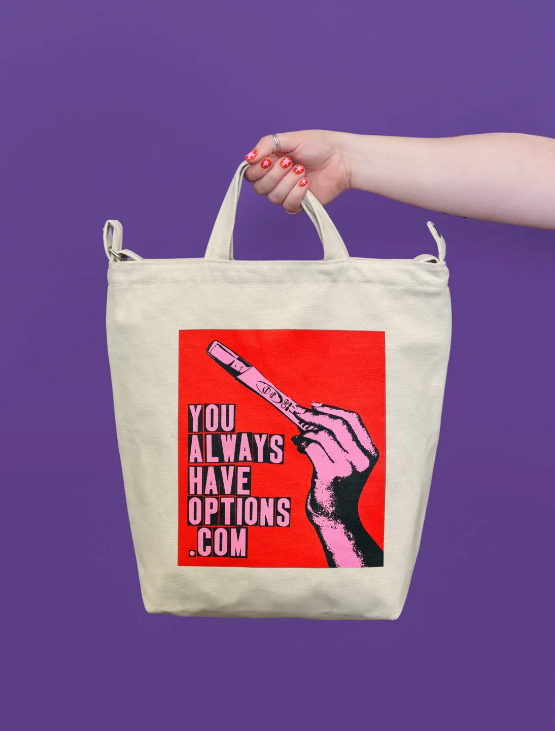 Light-skinned arm with red nails holding a natural canvas tote bag with a red, blank and pink graphic that says YOU ALWAYS HAVE OPTIONS .COM