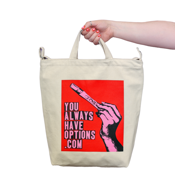 Light-skinned arm with red nails holding a natural canvas tote bag with a red, blank and pink graphic that says YOU ALWAYS HAVE OPTIONS .COM