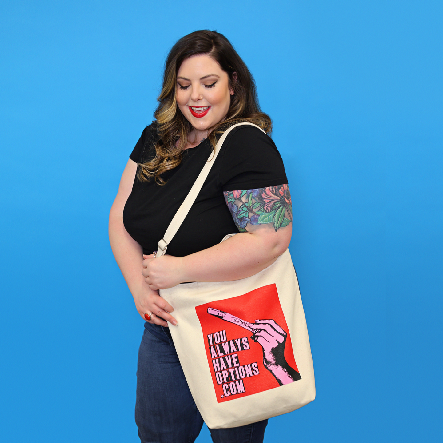 Femme person dressed in black, wearing a natural canvas tote bag with a red, blank and pink graphic that says YOU ALWAYS HAVE OPTIONS .COM