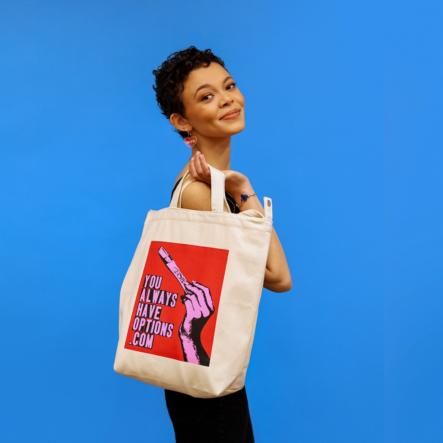 Femme person dressed in black, wearing a natural canvas tote bag with a red, blank and pink graphic that says YOU ALWAYS HAVE OPTIONS .COM