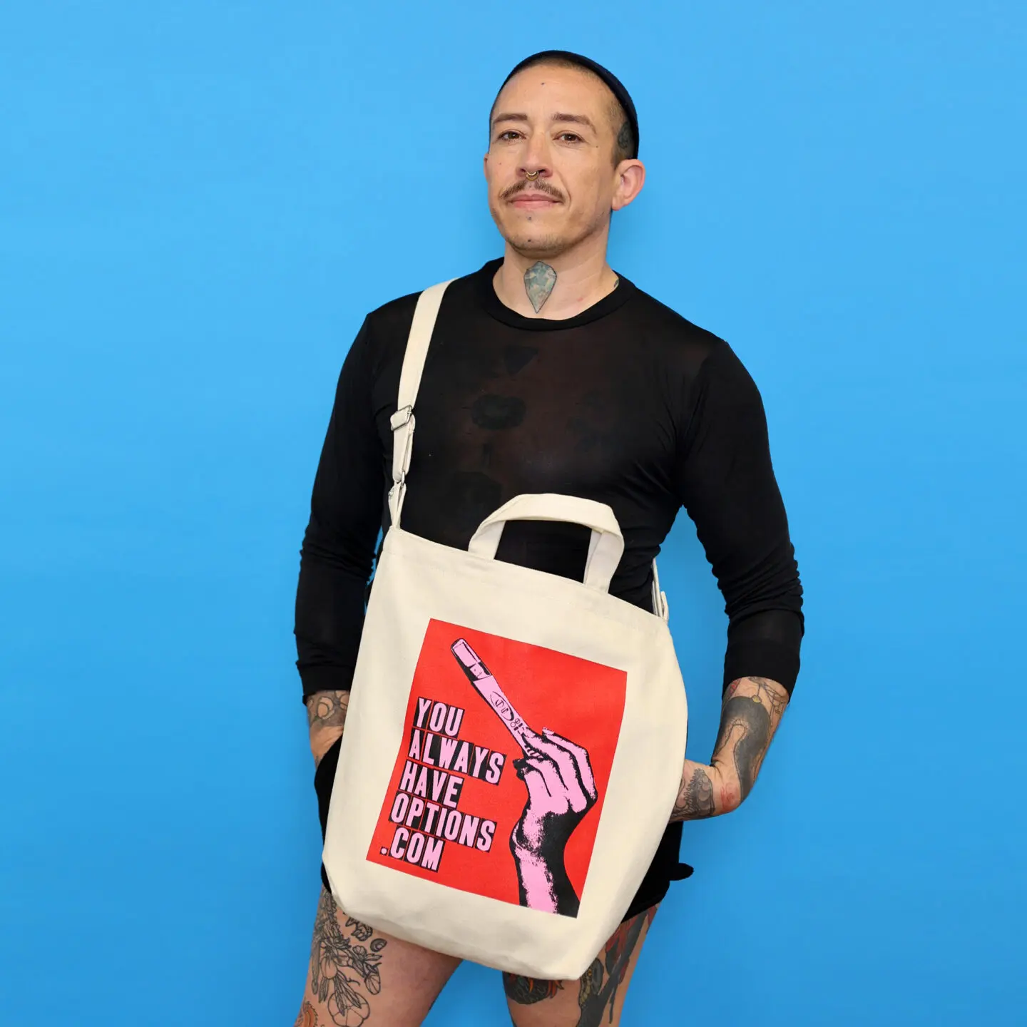 Masc person dressed in black, wearing a natural canvas tote bag with a red, blank and pink graphic that says YOU ALWAYS HAVE OPTIONS .COM
