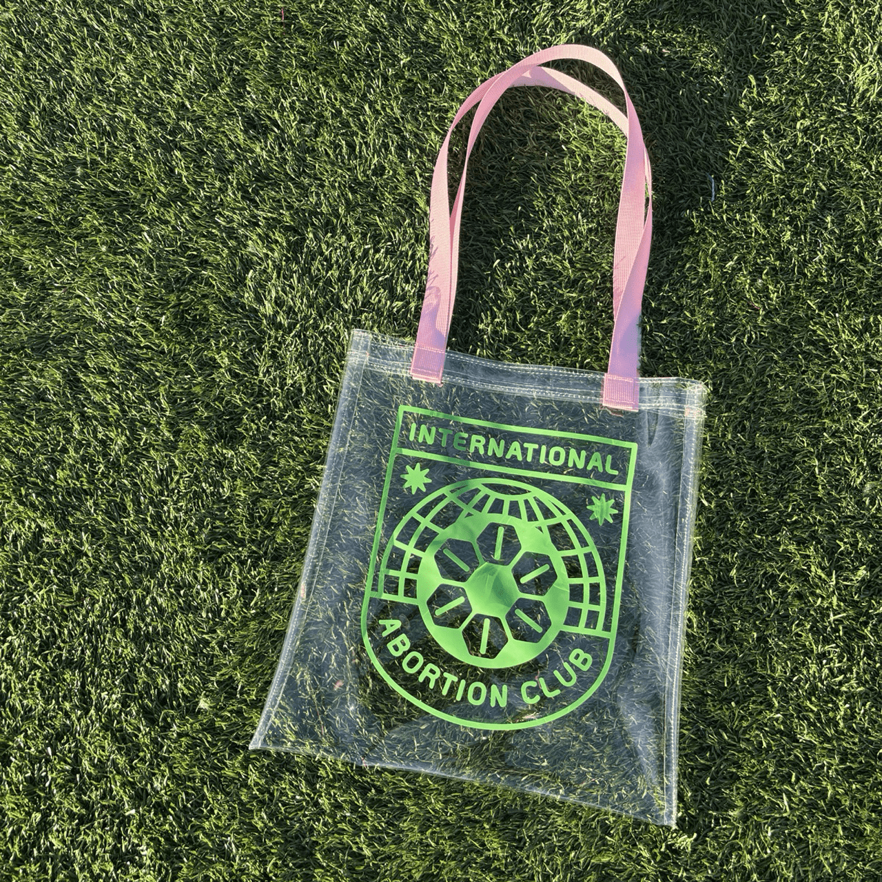 Clear vinyl tote with pink handles and the International Abortion Club logo in kelly green on one side, laying on grass